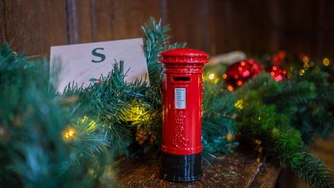 A small red postbox on a wooden bench with green foliage around it, and an envelope next to it with the letter S on.